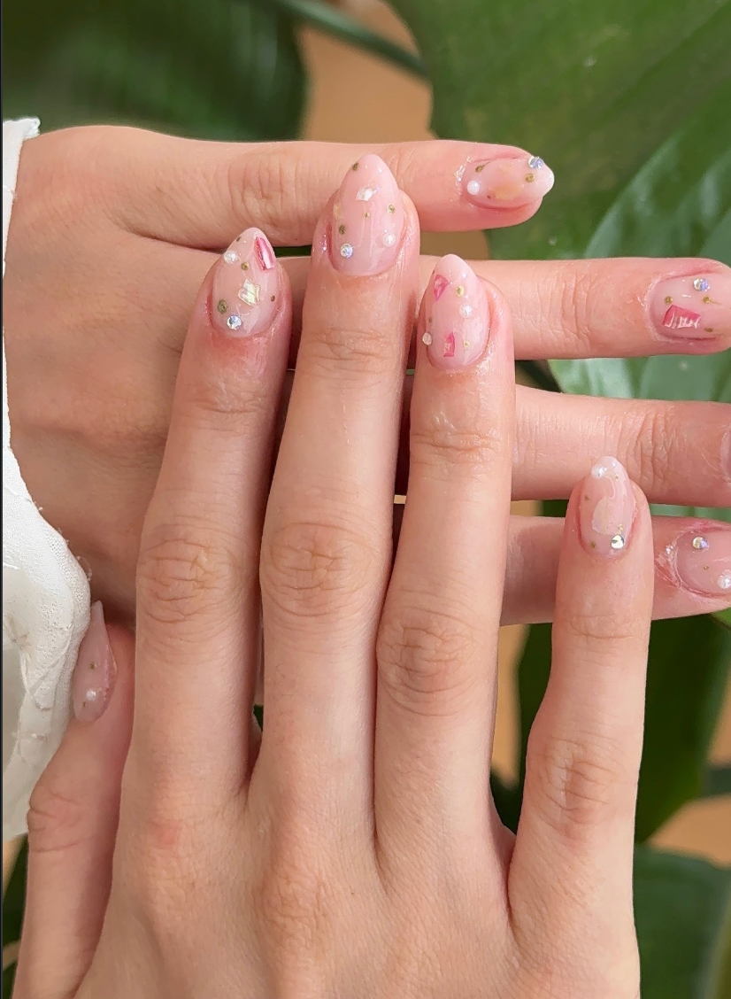 Uñas con diseño minimalista de pedrería pequeña sobre esmalte nude, en primer plano sobre un fondo verde difuminado