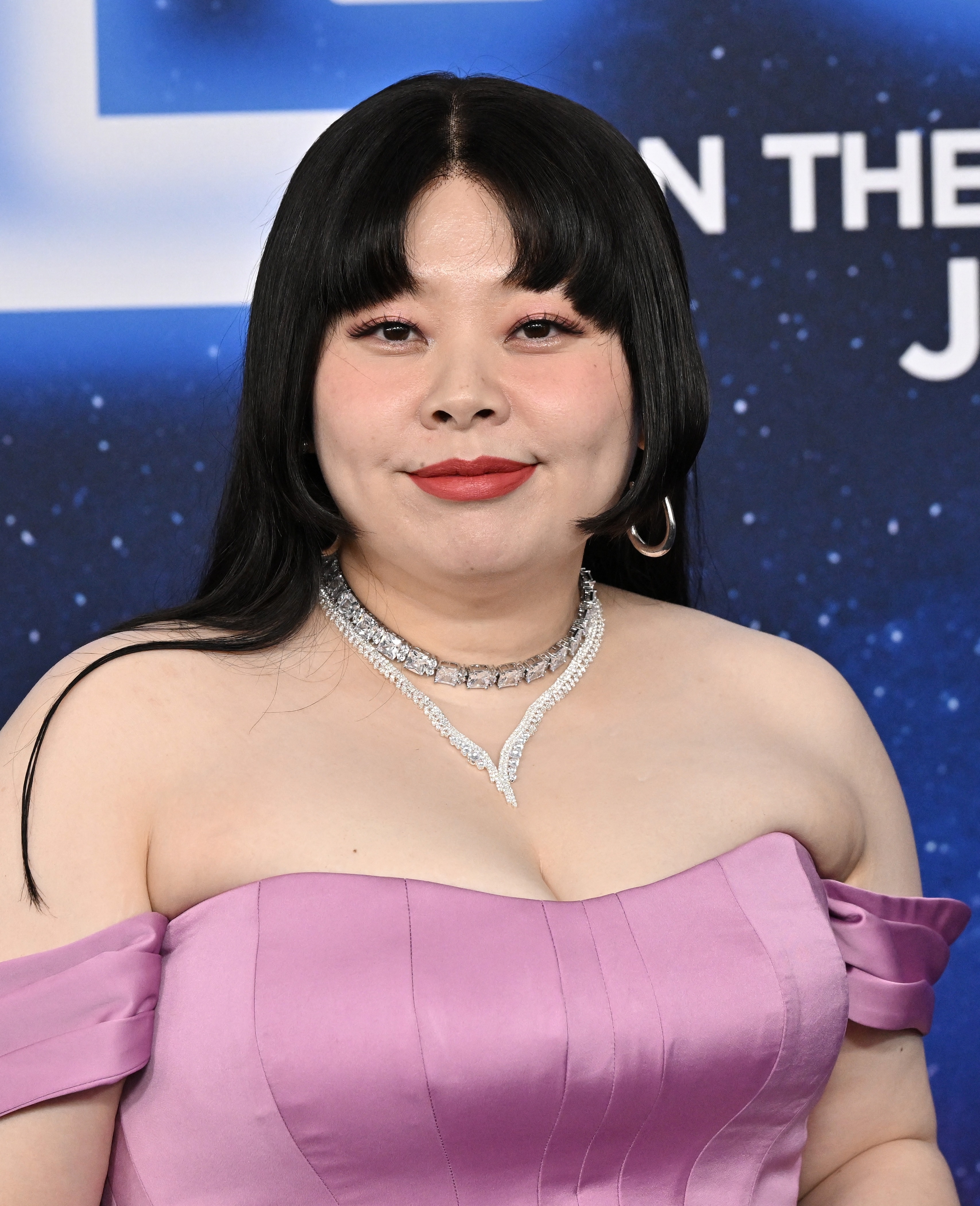 女性がパープルのオフショルダードレスと輝く宝石を身につけて微笑んでいる。背景にはイベントの一部が見える。