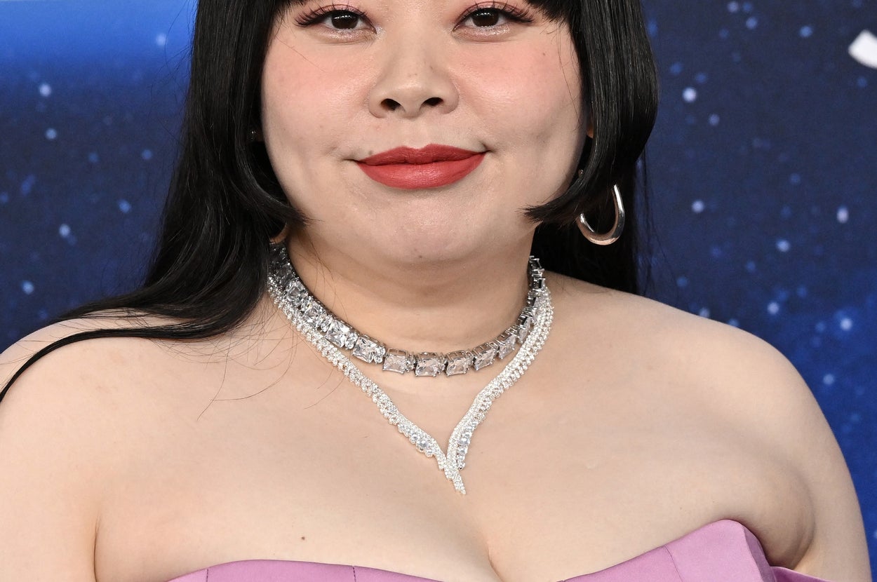 女性がパープルのオフショルダードレスと輝く宝石を身につけて微笑んでいる。背景にはイベントの一部が見える。