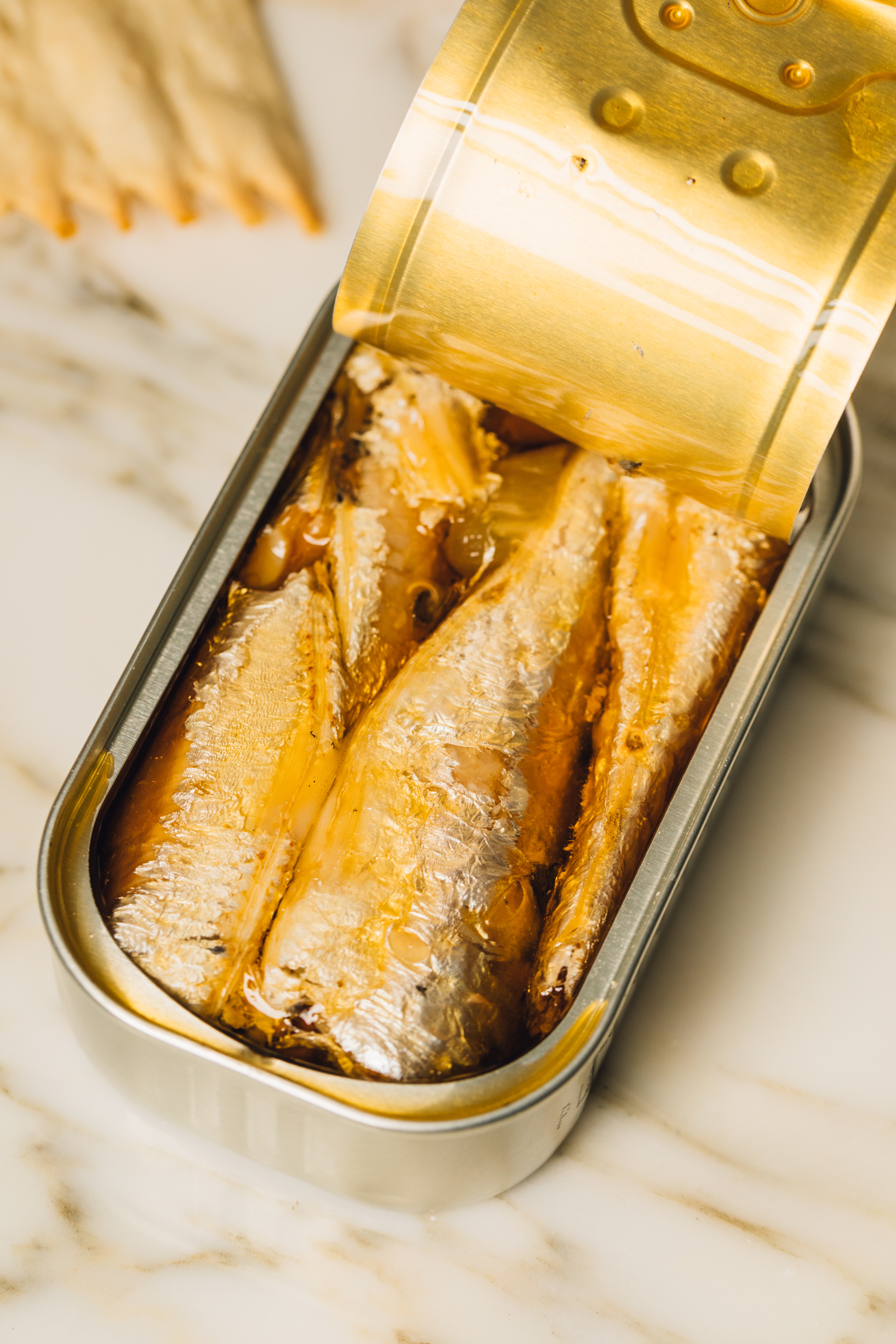 Open can of sardines on a marble surface, with some crackers in the background