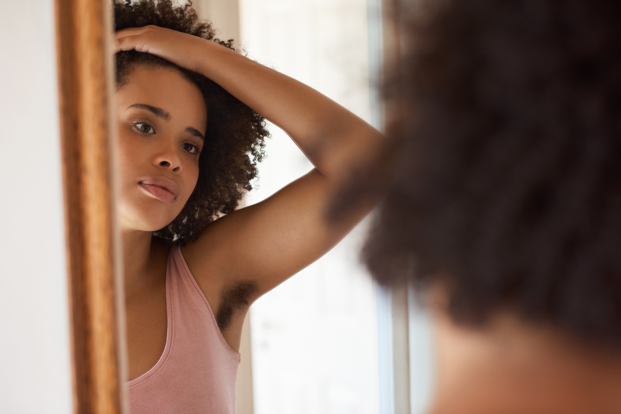 Osoba o kręconych włosach patrząca w lustro, ubrana w top bez rękawów, z kontemplacyjnym wyrazem twarzy