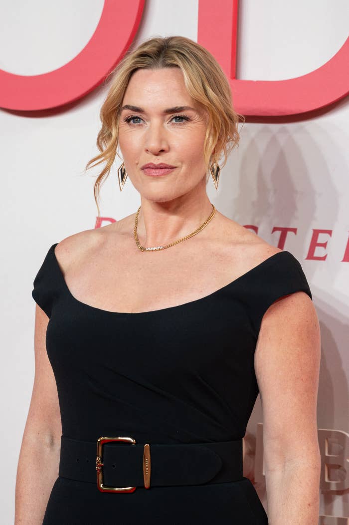 Person in elegant off-the-shoulder dress on red carpet, wearing statement earrings and necklace, with hair styled in loose waves