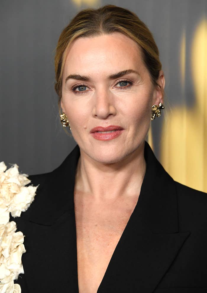 A person in a black suit jacket with floral details and gold earrings, posing for a portrait