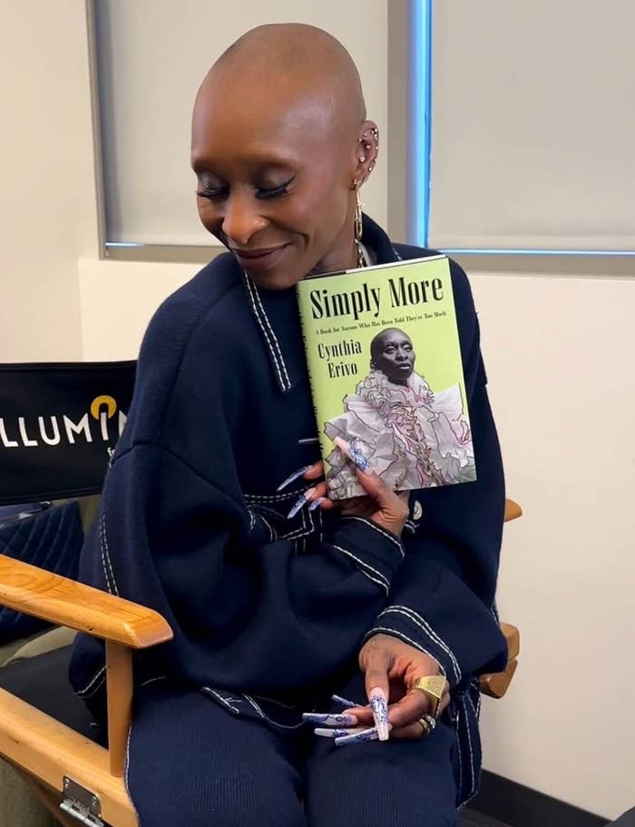 Person with shaved head, wearing a chic outfit, holds up a book titled &quot;Simply More&quot; and smiles in a seated setting