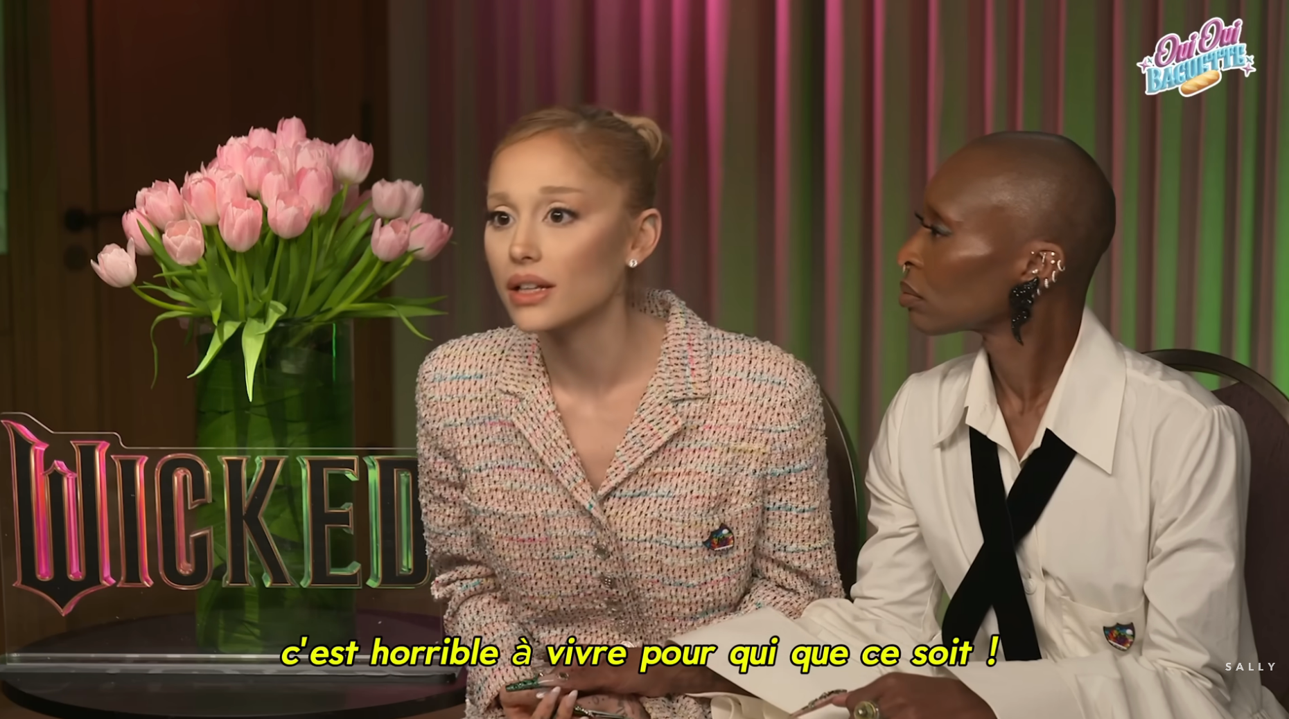 Two individuals discuss in an interview setting, with a bouquet of tulips and &quot;Wicked&quot; sign visible. French subtitle shown