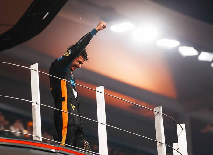 Race car driver celebrates on a podium, raising a fist triumphantly, wearing a racing suit, with a crowd in the background