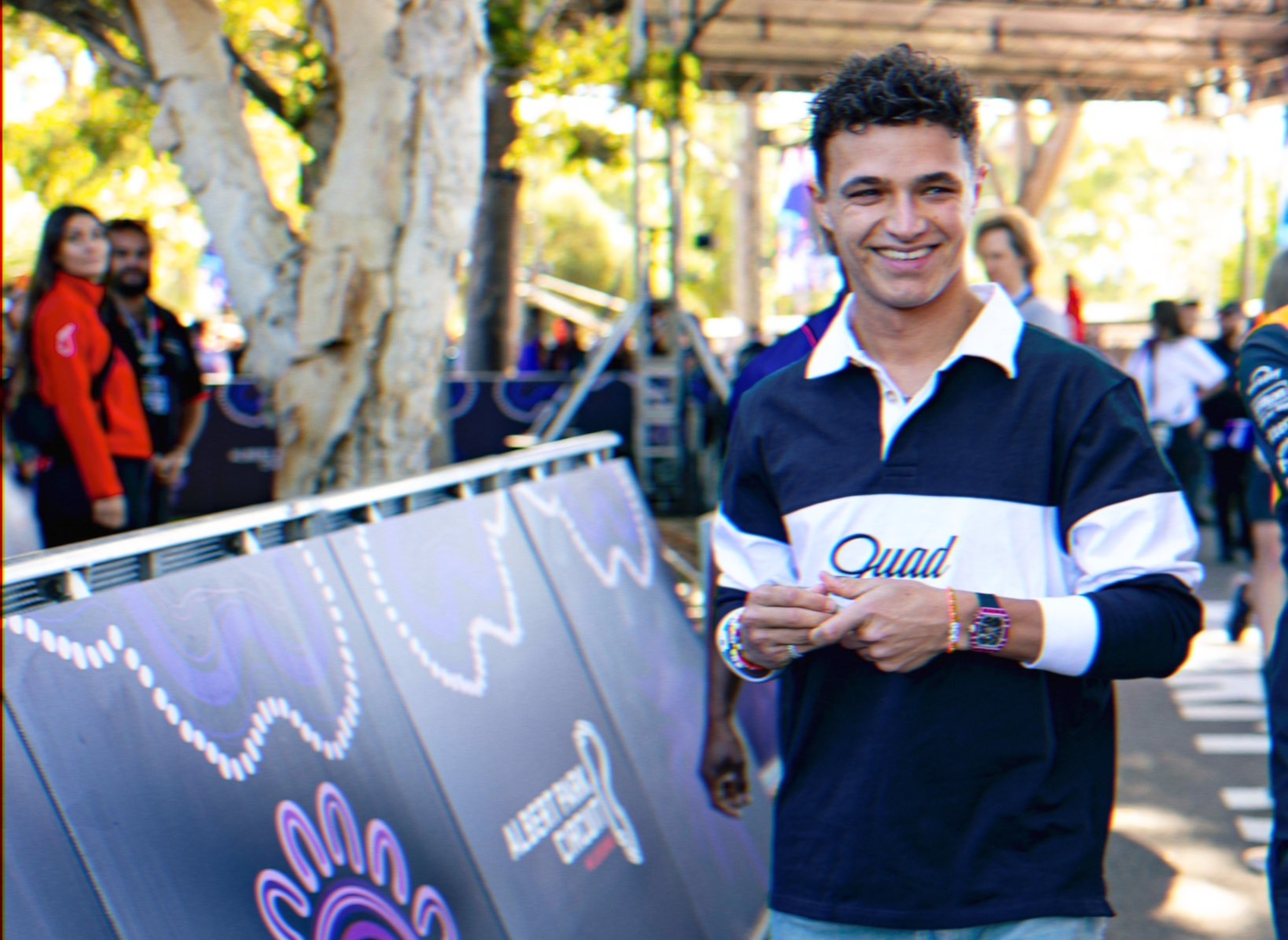 Person smiling, wears casual long-sleeve polo and jeans, stands near a barricade at a public event. Trees and banners visible in the background