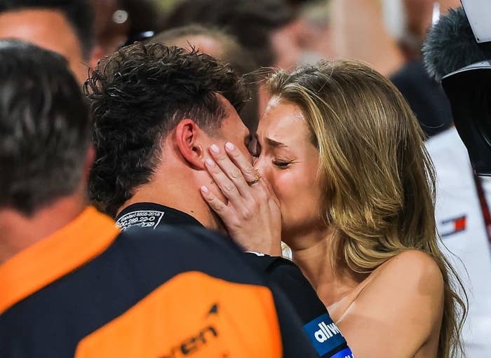Woman kisses a man in a McLaren race suit amidst a group of people, with media capturing the moment