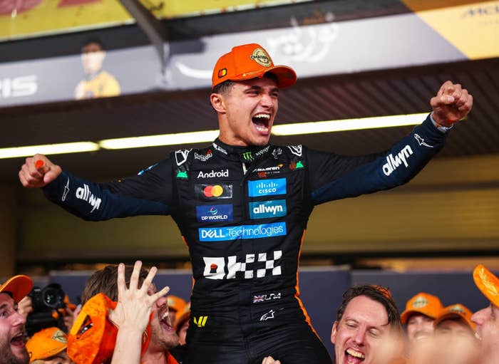 Race car driver in a sponsor-branded jumpsuit, jubilantly celebrating with a group while wearing a cap, cheering and lifting his arms