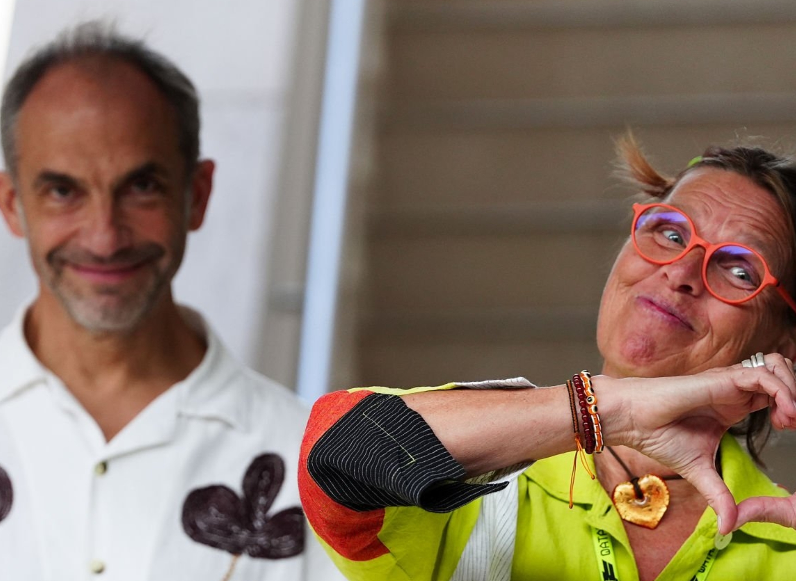 Two individuals pose, one wearing a white, floral-patterned shirt holding a soda can, another in vibrant attire with glasses, making a heart gesture