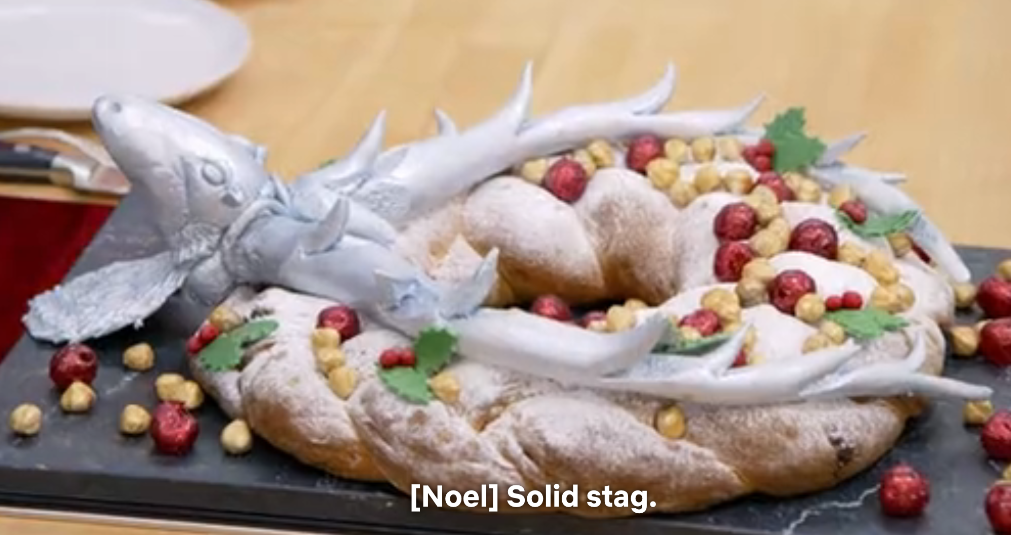 A festive bread centerpiece shaped like a stag, adorned with berries and nuts, dusted with powdered sugar. Subtitles: "[Noel] Solid stag."