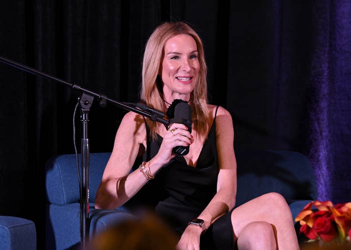 A person in a black dress sits on a stage, speaking into a microphone during an event or interview