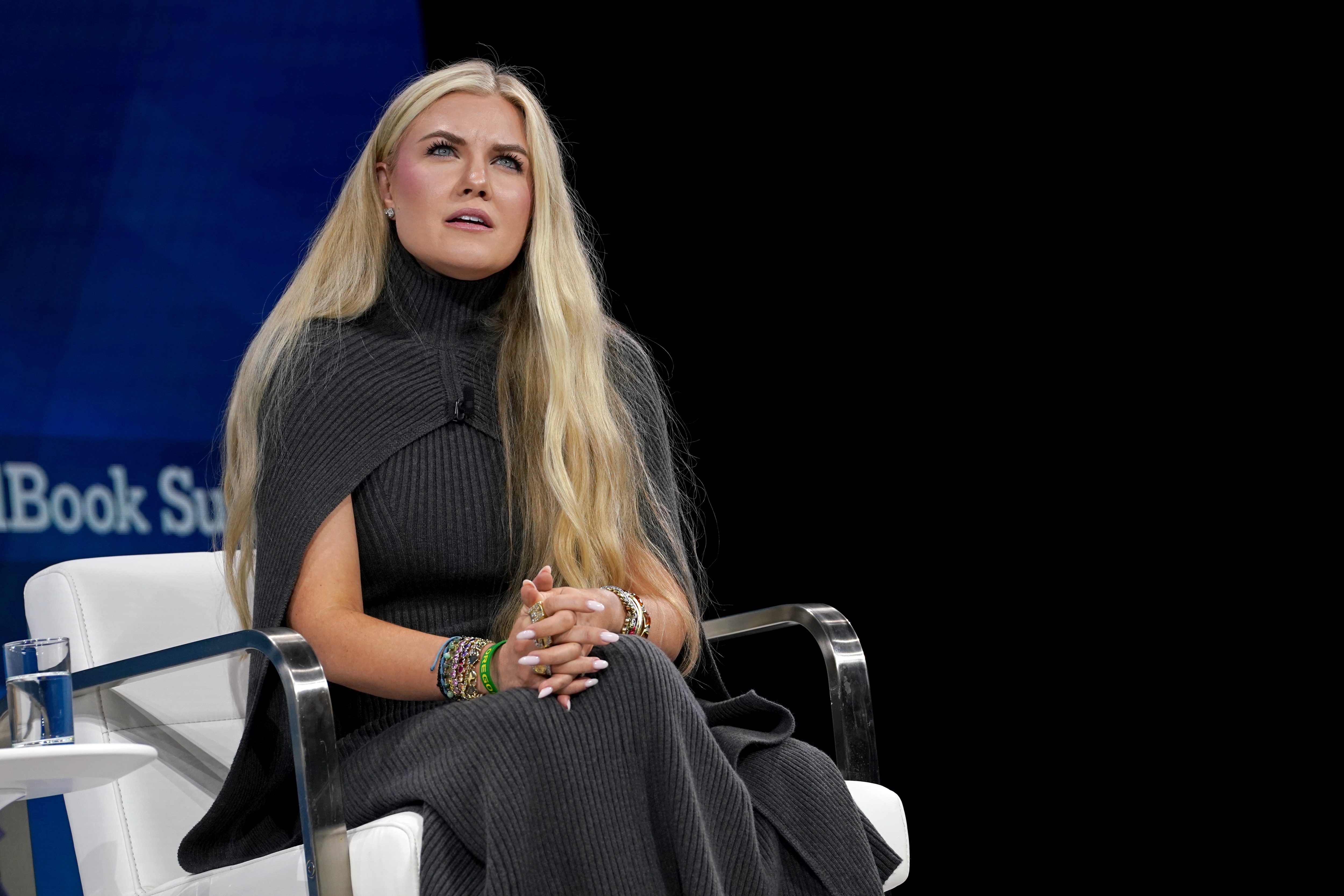 Person seated, wearing a ribbed knit dress with a cape-like top, speaking at an event
