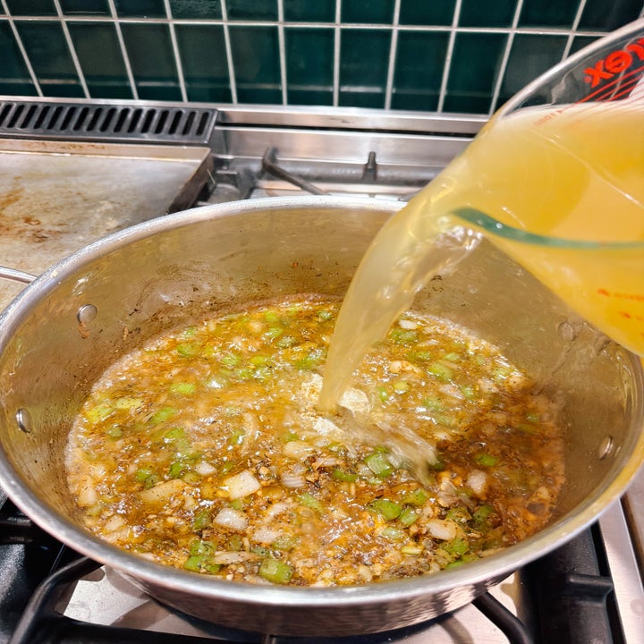 Broth being poured into a pot with diced onions and celery on a stove, likely in preparation for a soup or stew