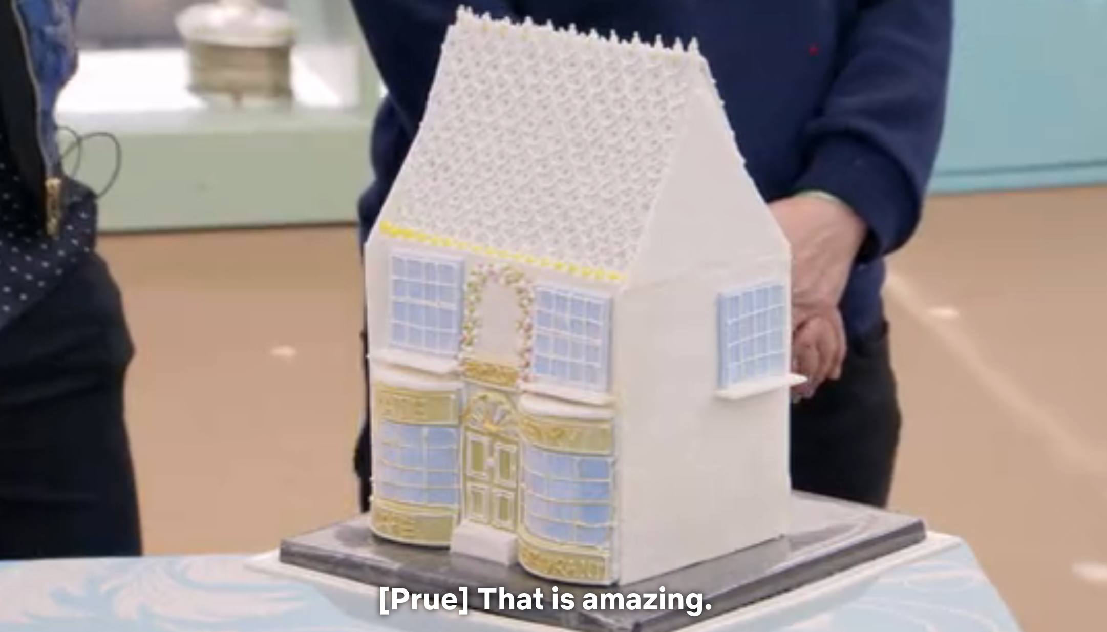 Elaborate cake shaped like a house with intricate details admired by two people, one saying, "That is amazing."