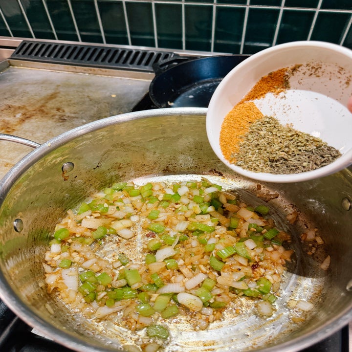 Chopped onions and celery sautéing in a pot, with a hand adding mixed spices from a small bowl above