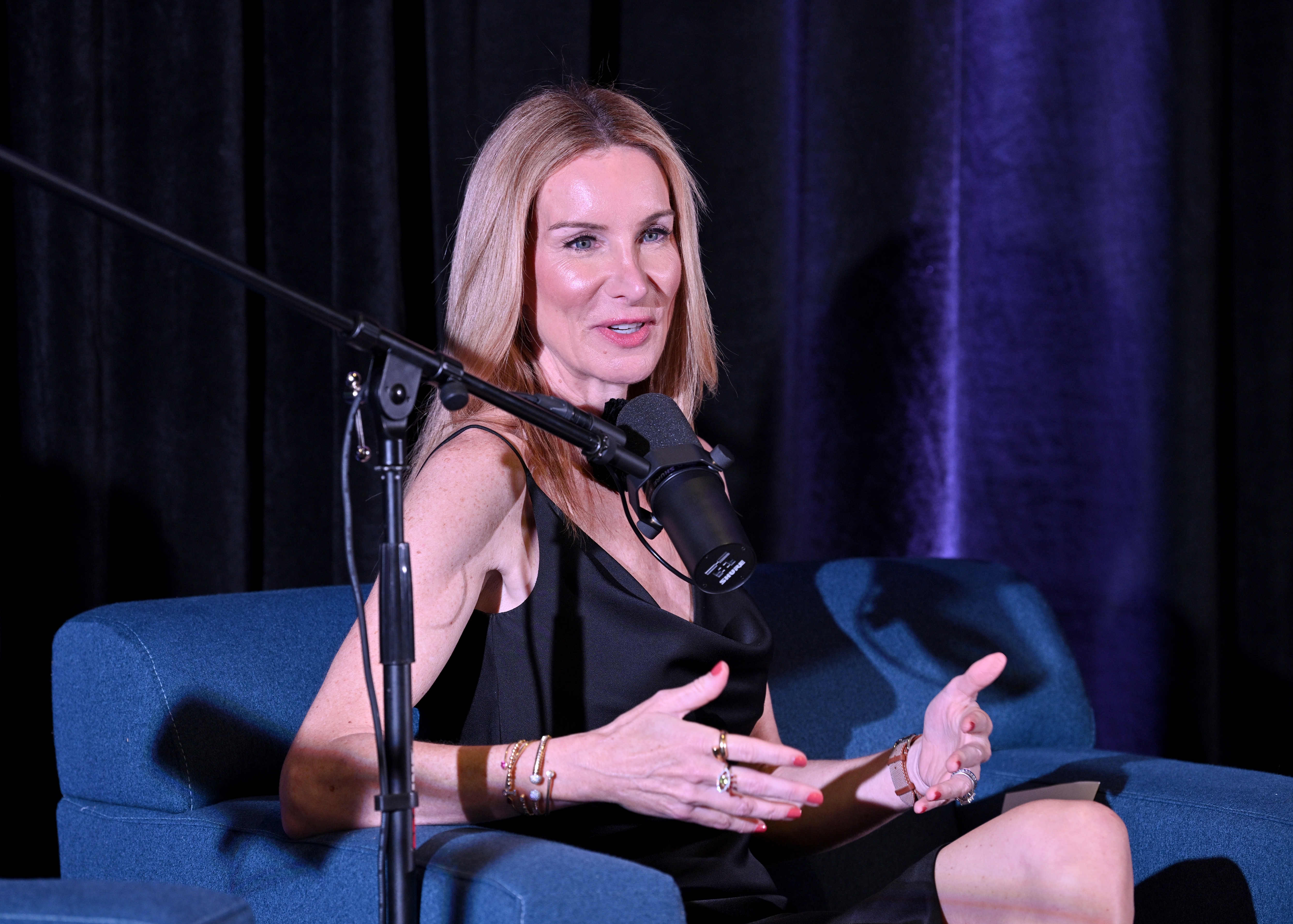 A person sits on a blue chair, wearing a black sleeveless dress, speaking into a microphone during an interview or talk event