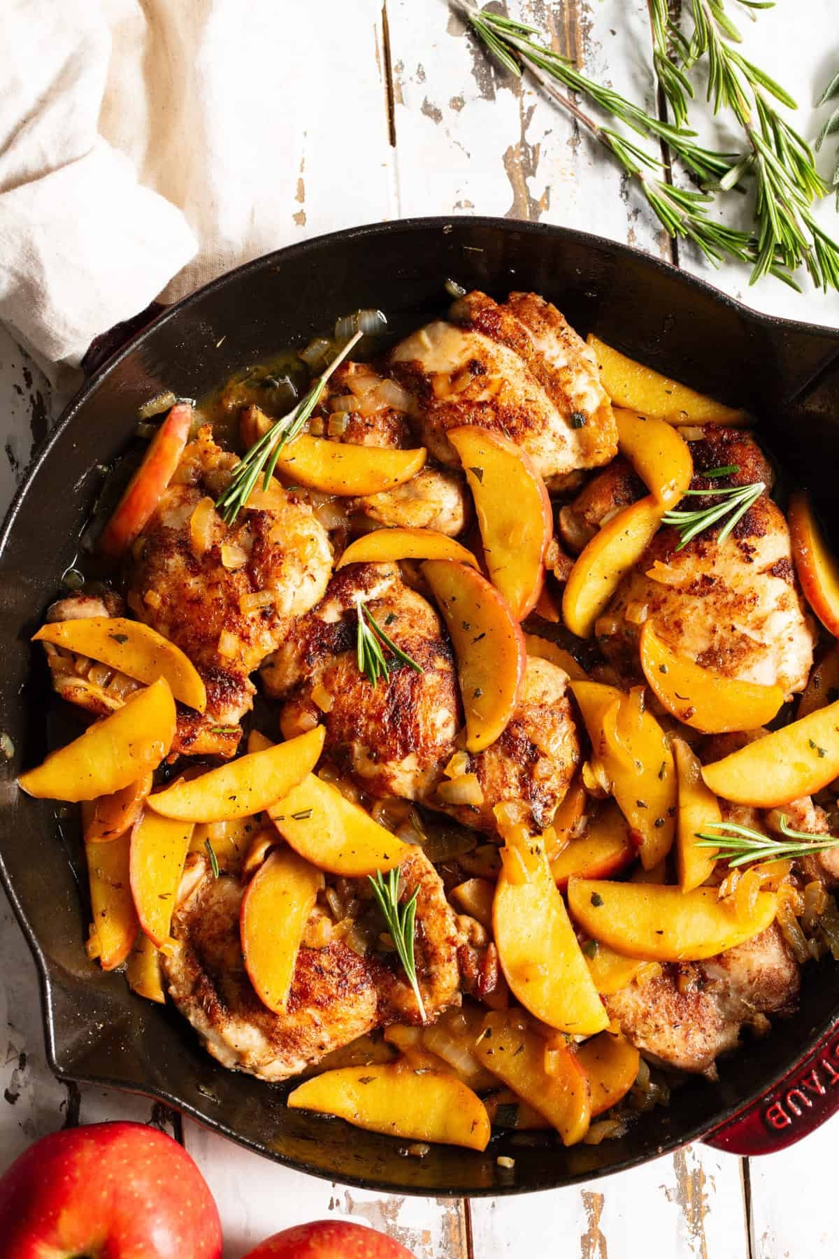 Skillet with cooked chicken thighs topped with slices of apples and rosemary sprigs, resting on a rustic surface with apples nearby