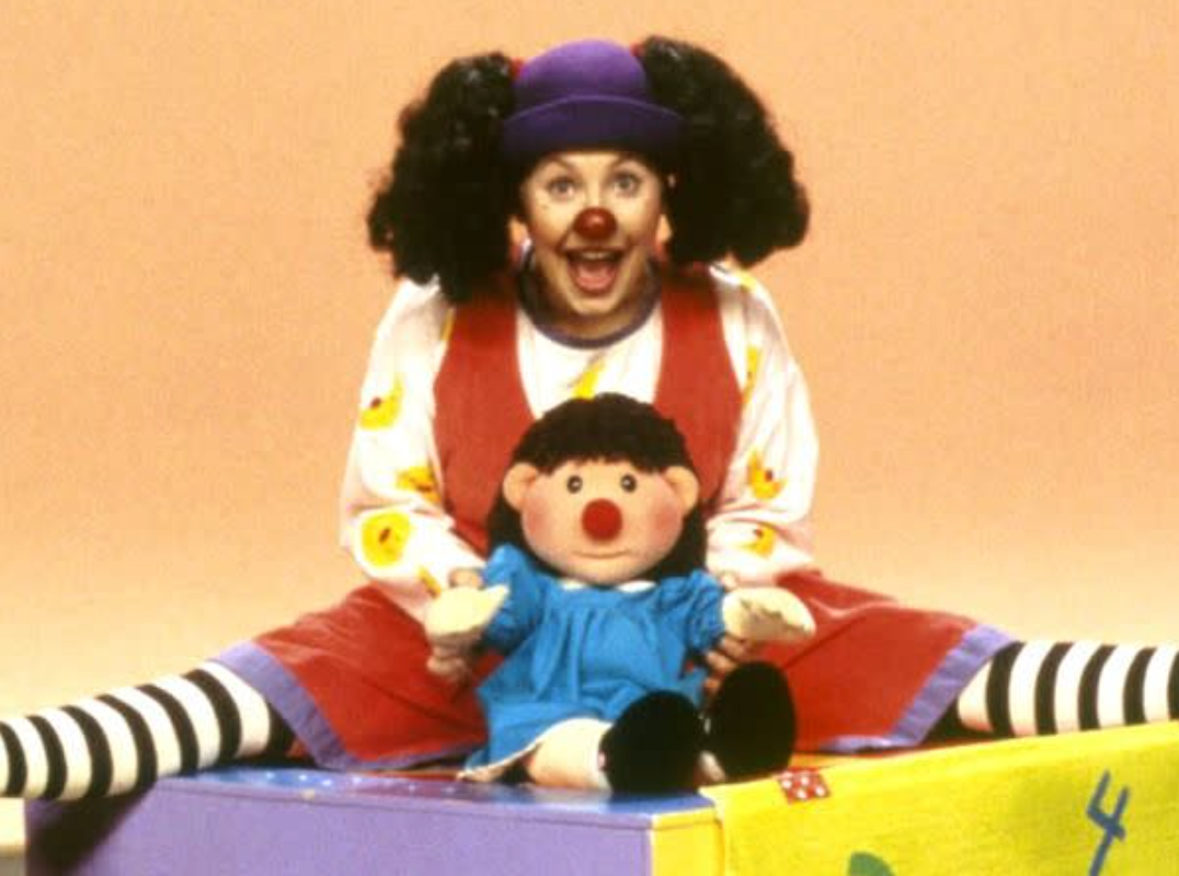 Person kledd som en klovn med lilla hatt og stripete sokker, sittende på en fargerik blokk og holder en dukke med lignende trekk