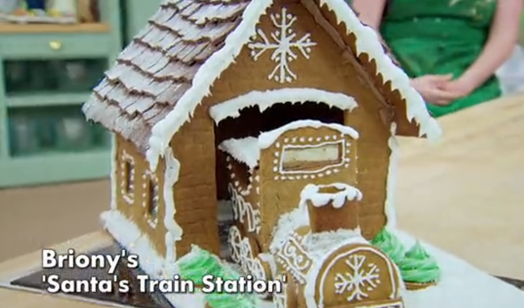 Gingerbread train station decoration with icing details and text reading "Briony's 'Santa's Train Station'" on display in a kitchen setting