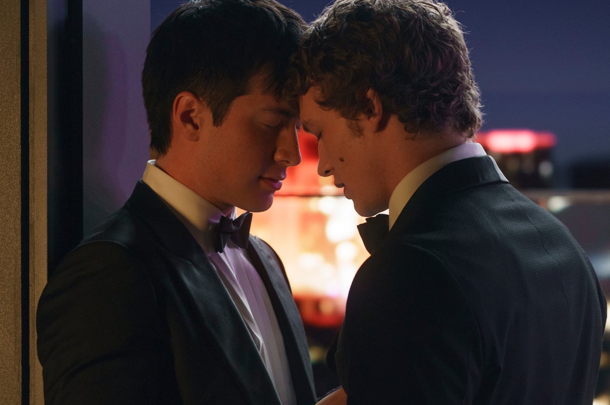 Two men in tuxedos share an intimate moment, leaning against a wall in a softly lit setting, suggesting a romantic scene from a TV or movie