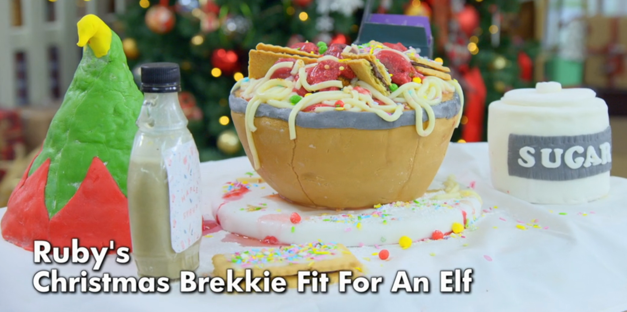 A whimsical breakfast-themed cake with spaghetti and sauce topping, labeled "Ruby's Christmas Brekkie Fit For An Elf," beside a bottle and sugar jar
