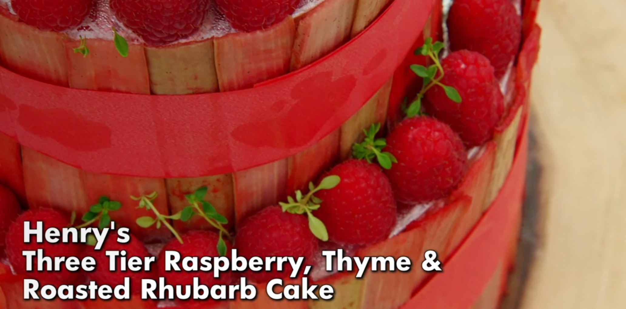 Three-tier cake with raspberries and roasted rhubarb, adorned with fresh thyme. Visible text: "Henry's Three Tier Raspberry, Thyme &amp;amp; Roasted Rhubarb Cake."