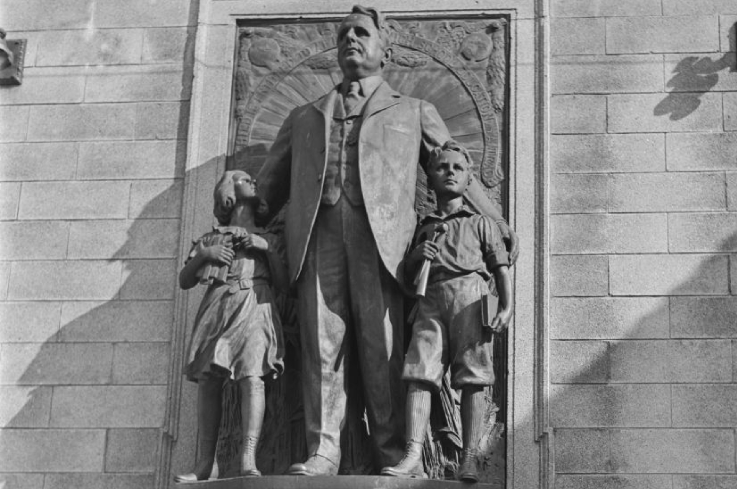 En bronsestatue av en mann i dress med to barn, en jente og en gutt, stående på en steinsokkel mot en dekorativ veggbakgrunn