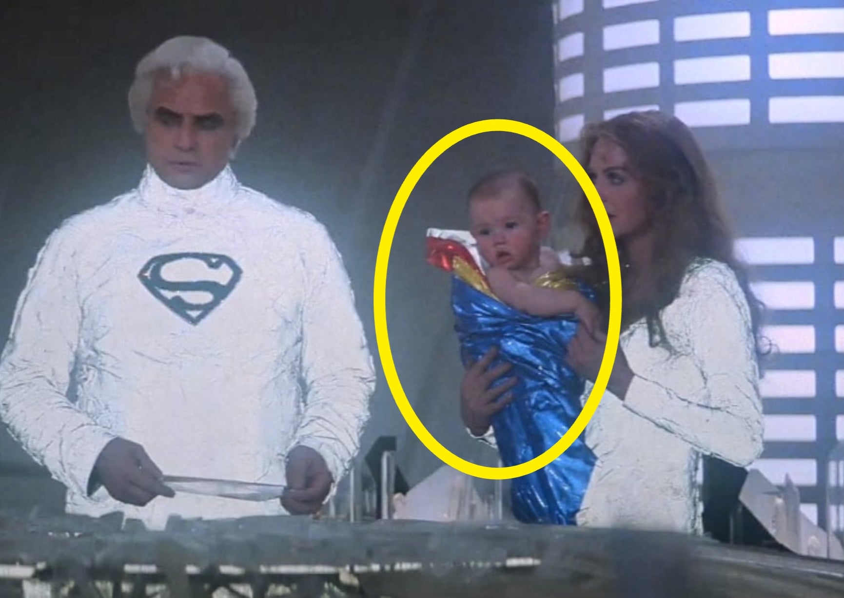 En mann og kvinne i futuristiske hvite antrekk holder en baby pakket inn i en superheltkappe, satt i et sci-fi-miljø