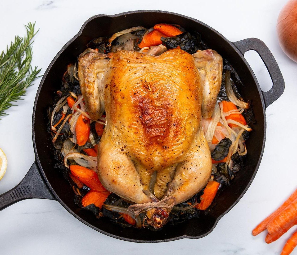 Roasted chicken in a skillet with sliced onions, carrots, and herbs. Lemons, rosemary, onions, and carrots on the side