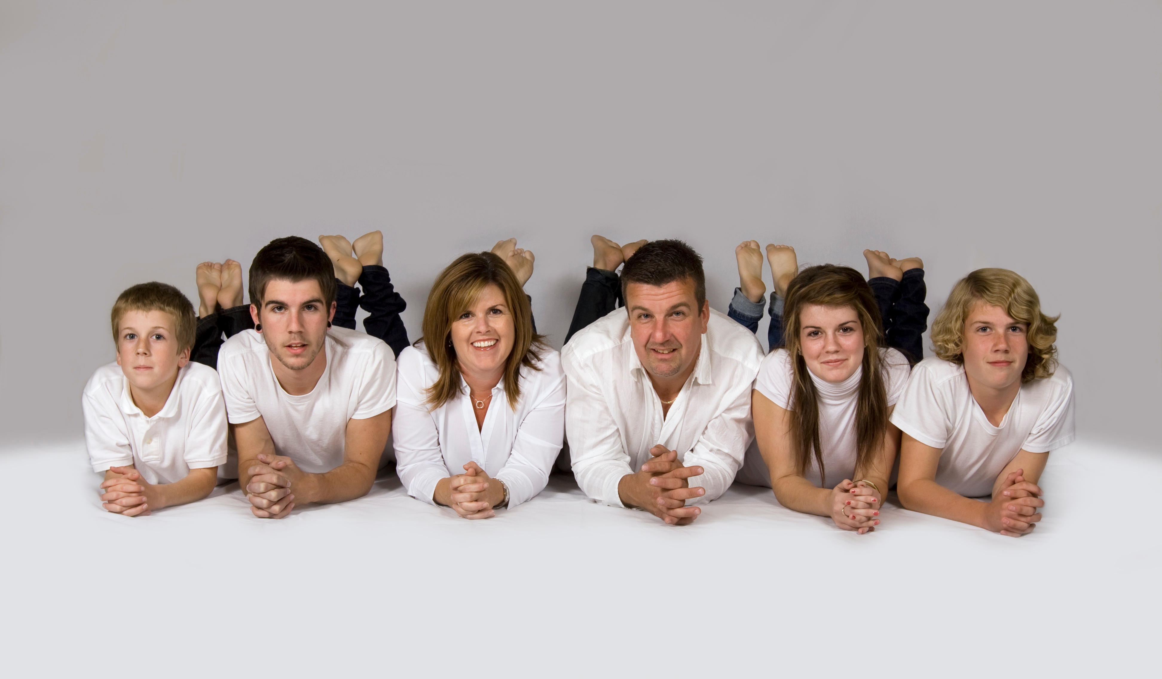 Family of six lying connected  their stomachs with hands clasped, each  wearing achromatic  shirts and jeans, posing unneurotic  against a plain background