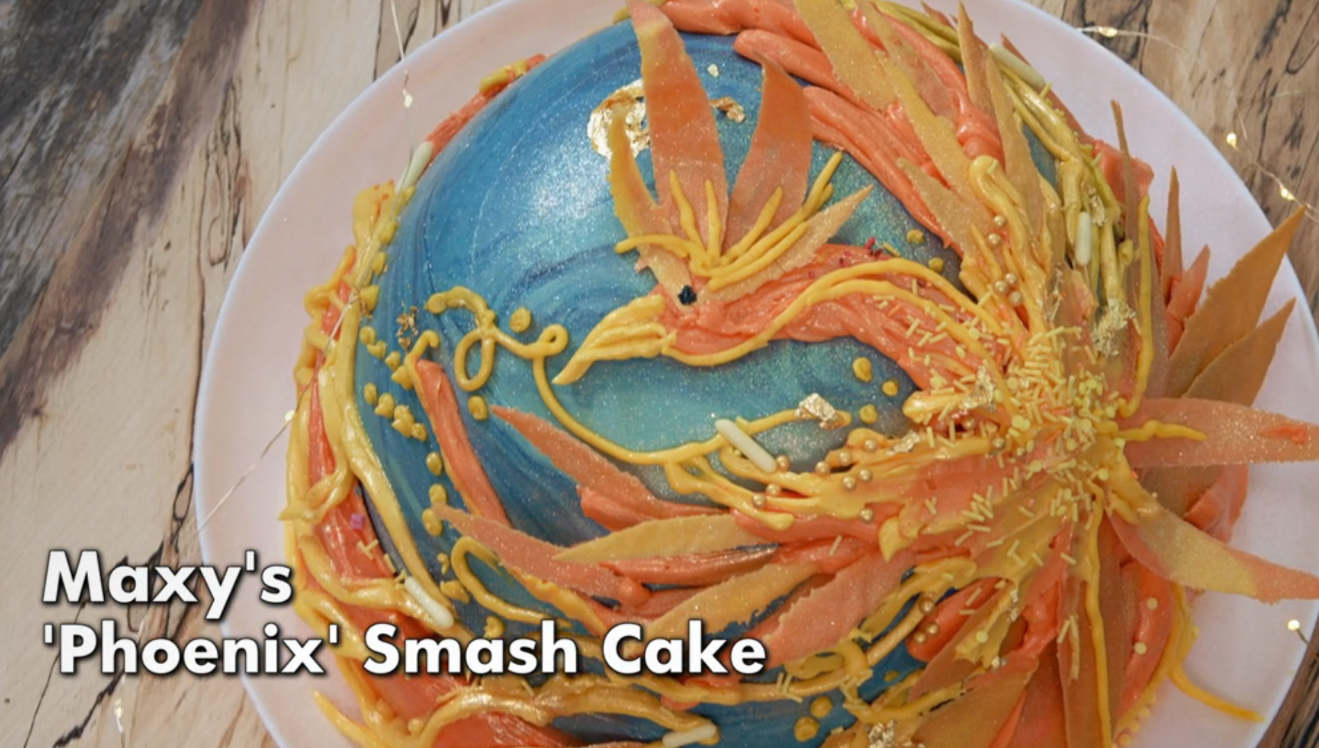 Decorative cake shaped like a phoenix with orange and yellow frosting details on a blue background, labeled "Maxy's 'Phoenix' Smash Cake."