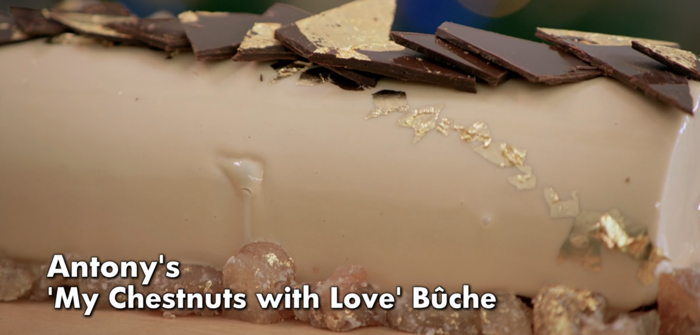 A log-shaped dessert titled "My Chestnuts with Love" Bûche, topped with gold leaf and chocolate shards, rests on a bed of candied chestnuts