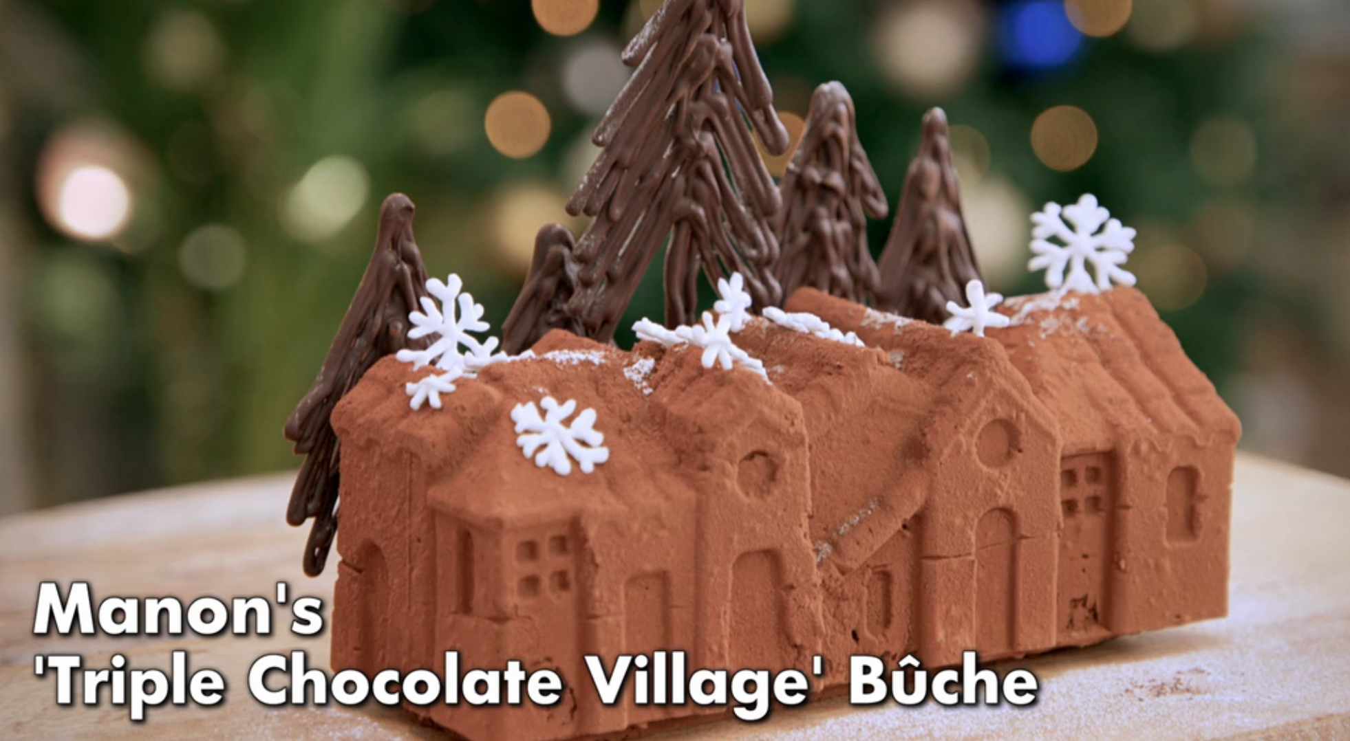 Chocolate village cake with tree decorations and snowflake accents, titled "Manon's 'Triple Chocolate Village' Bûche."