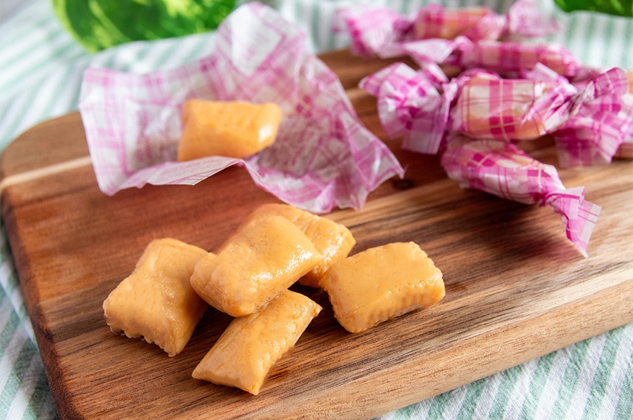 木のボードにキャラメルキャンディが並んでおり、奥には包み紙付きのキャンディが置かれている。背景に緑の葉。