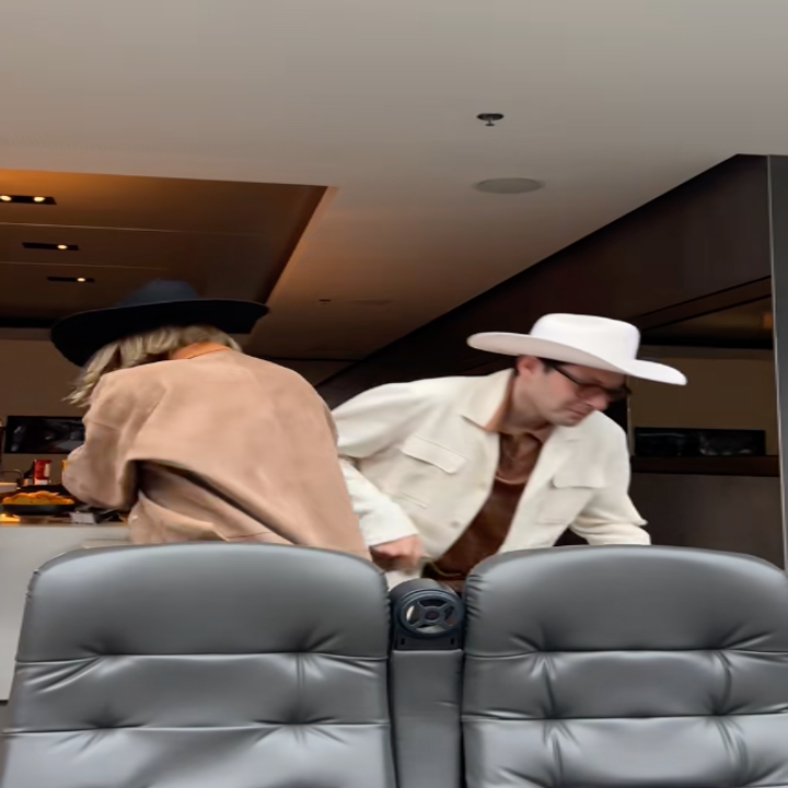 Two people wearing cowboy hats in a modern indoor setting, one in a tan jacket, the other in a white jacket, both facing away