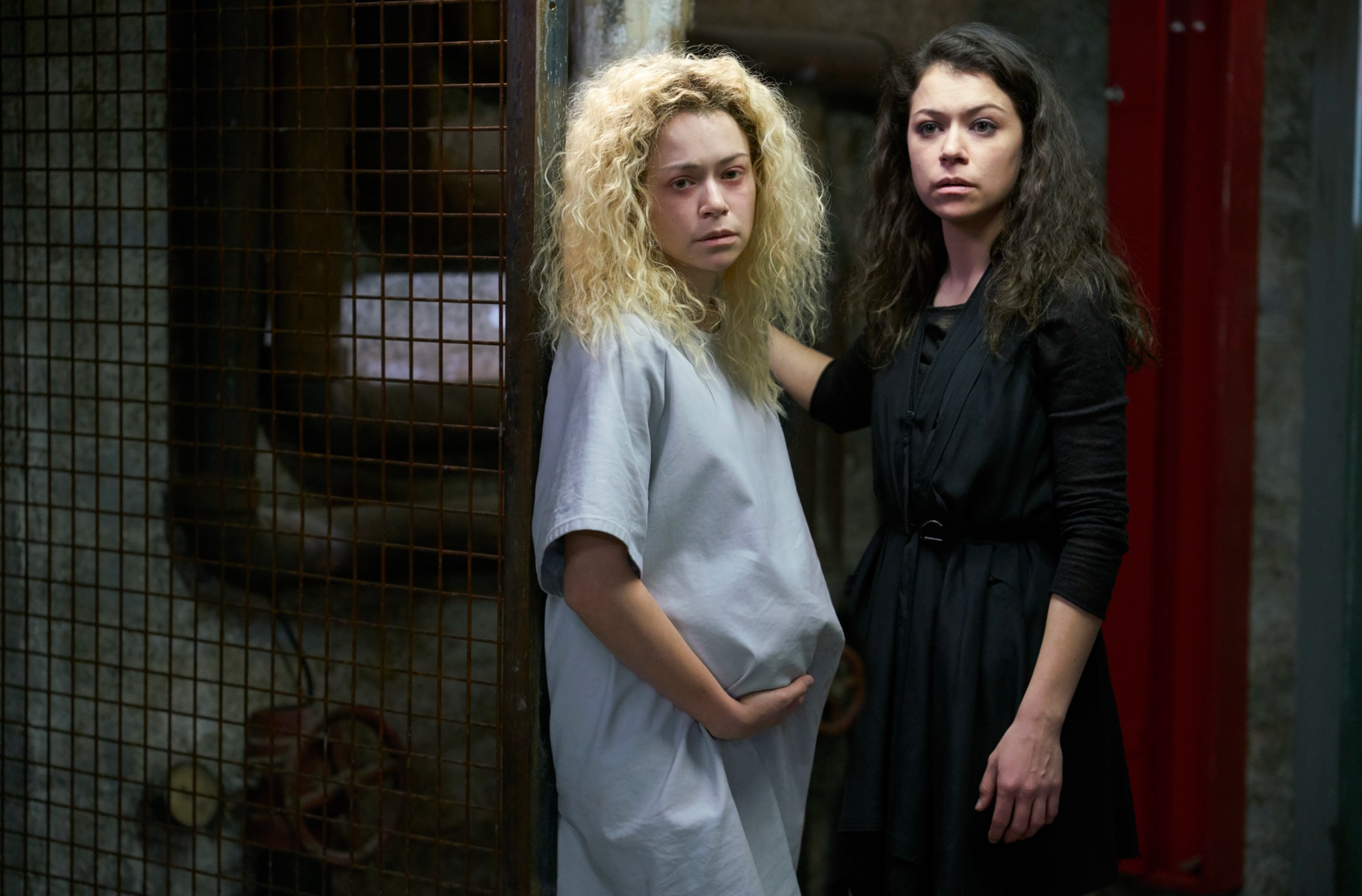 To kvinner står i en svakt opplyst setting, den ene med vilt krøllete hår i en sykehuskjole, den andre i en mørk, elegant kjole, og fremkaller en spent atmosfære