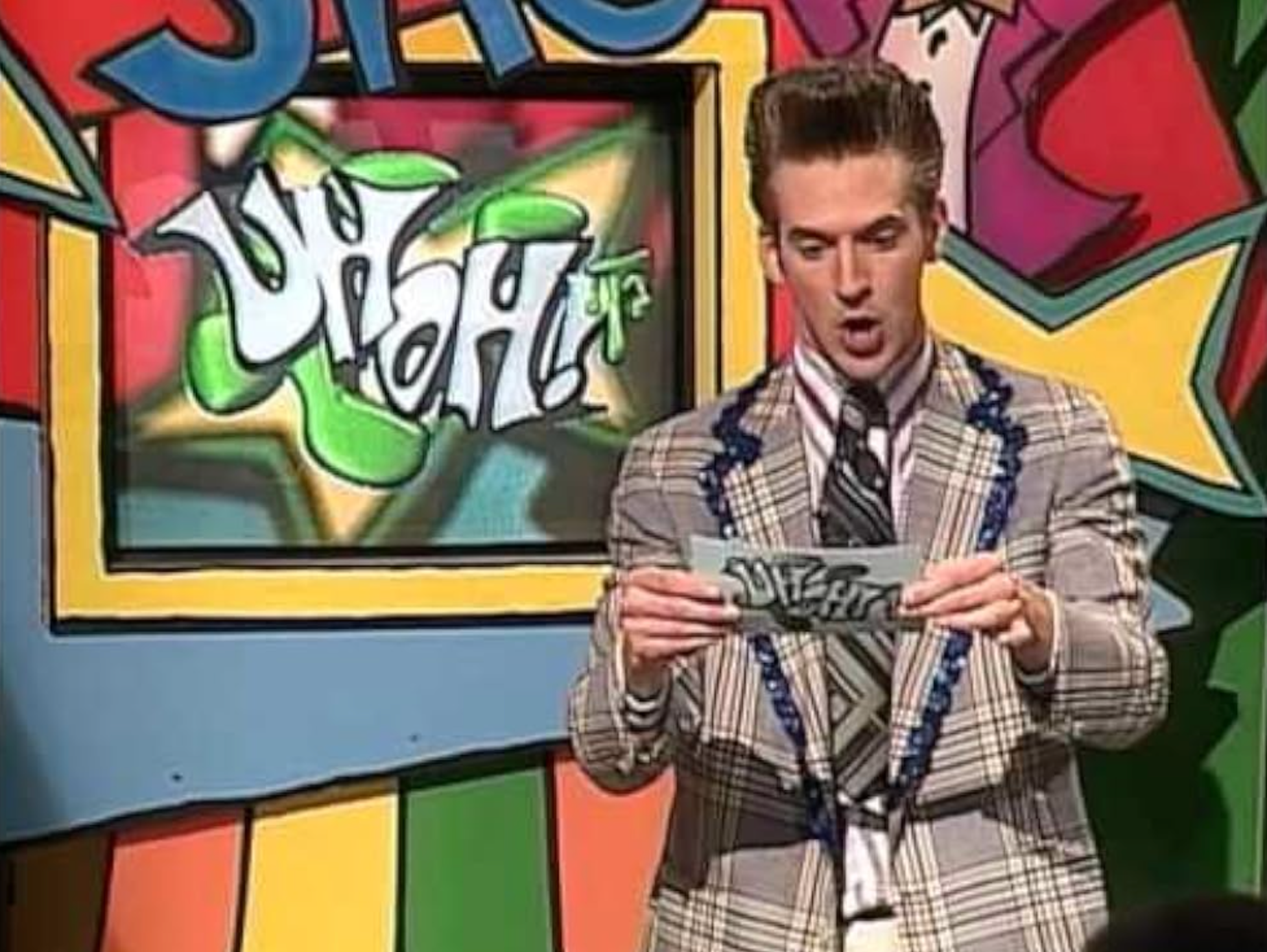 Person på et spillshow, holder et kort og presenterer. Skjermen bak viser ordet "Uh Oh!" Stilisert rutete dress med slips