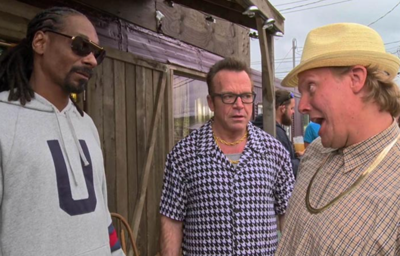 Tre menn snakker utendørs. En bruker solbriller og hettegenser, en annen har en mønstret skjorte, og den tredje har hatt og animert uttrykk