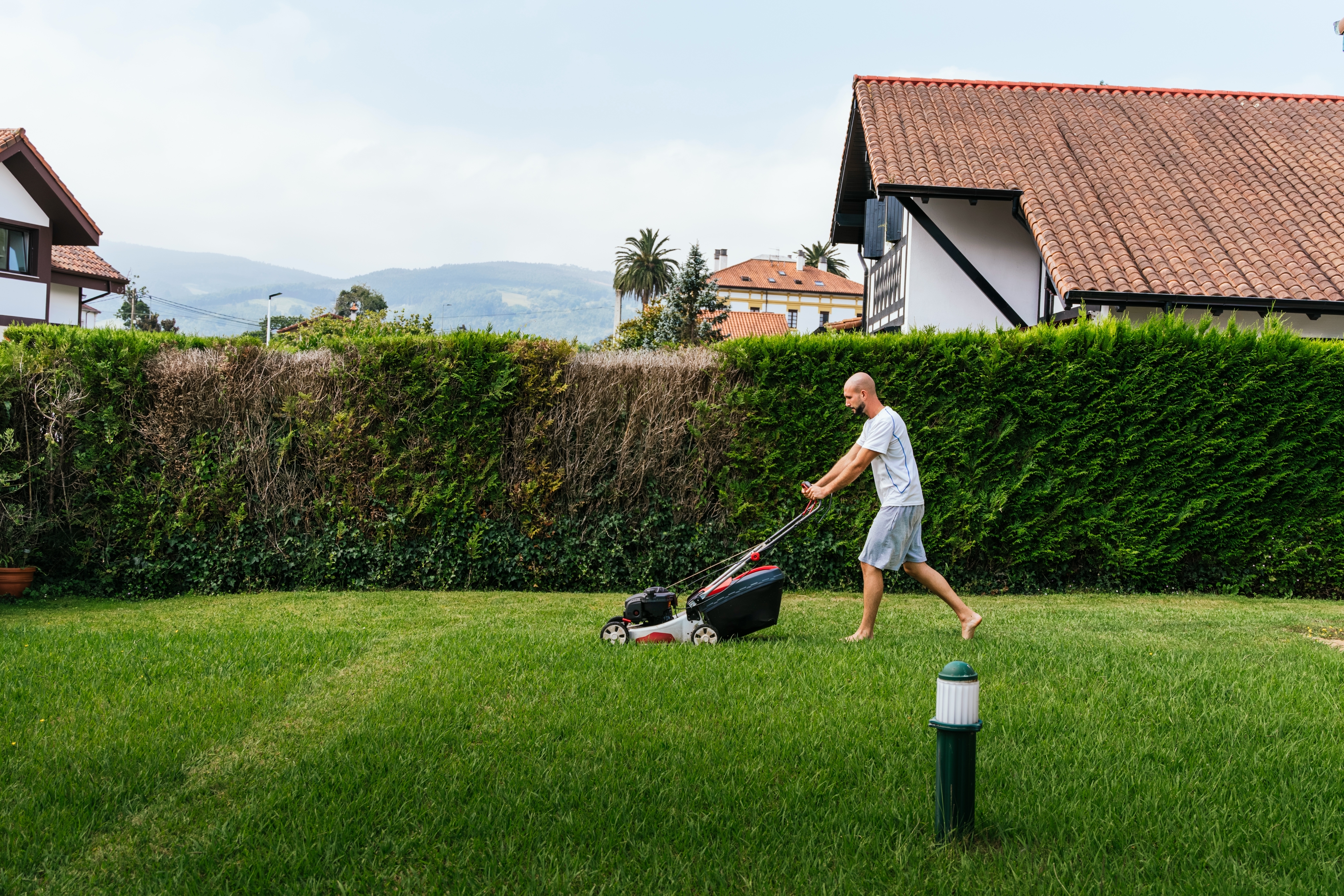 En person klipper en plen foran bolighus med fjell i bakgrunnen