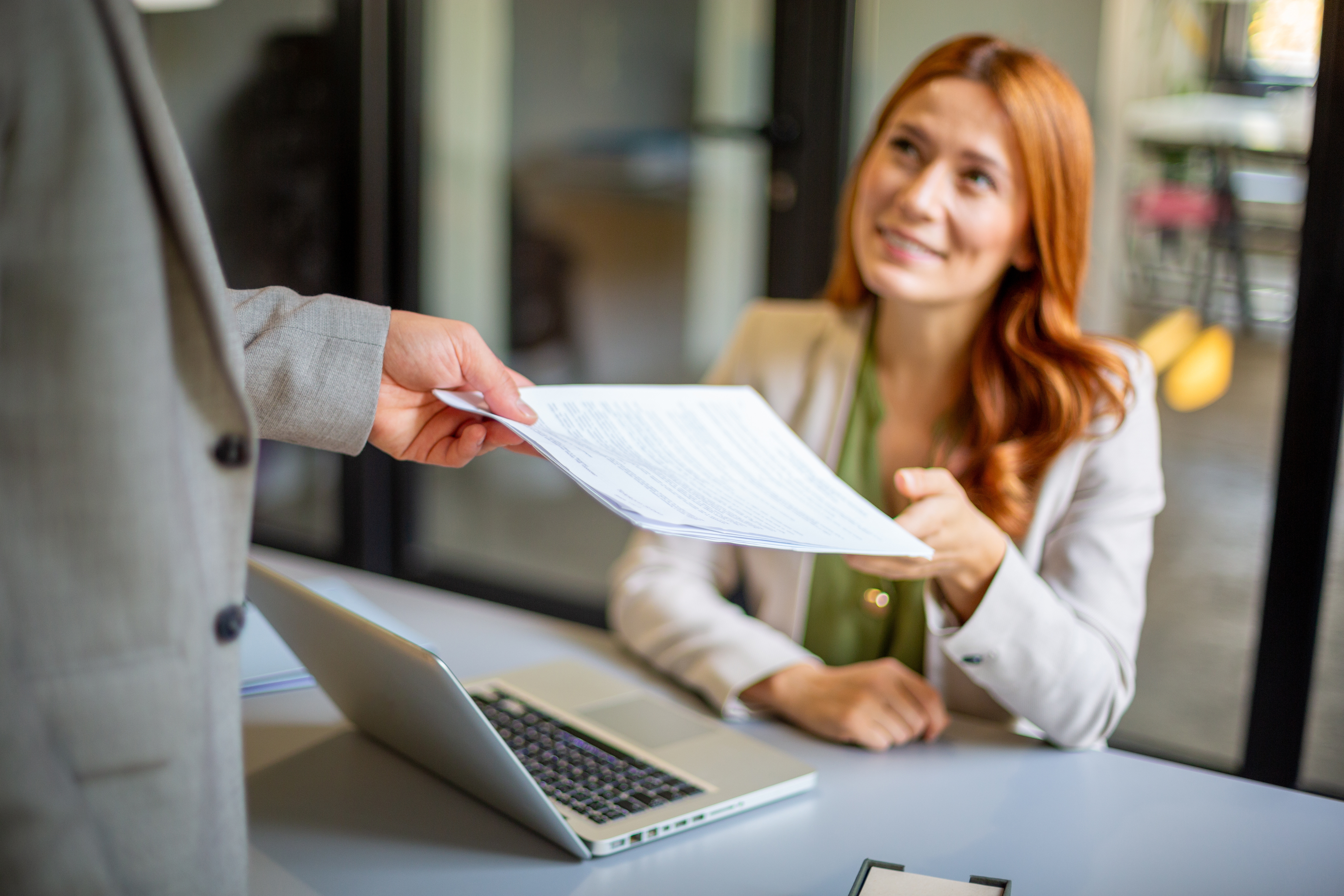 Person hands a papers  to a seated pistillate   successful  a nonrecreational  setting, with a laptop connected  the desk