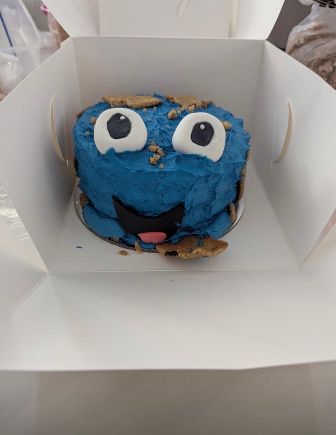 کیک دکور شده به شکل هیولای آبی با چشم‌های بزرگ و جزئیات کوکی درون یک جعبهٔ سفید