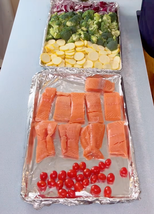 Salmon fillets and cherry tomatoes on one tray, broccoli, squash, and onions on another tray. Both trays are prepped, ready for baking