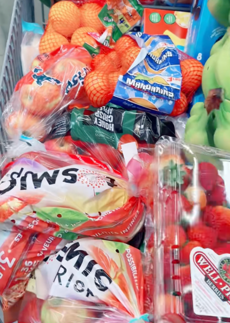 Shopping cart filled with packaged fruits like apples and strawberries, along with snacks such as macaroni and cheese boxes, in a grocery store