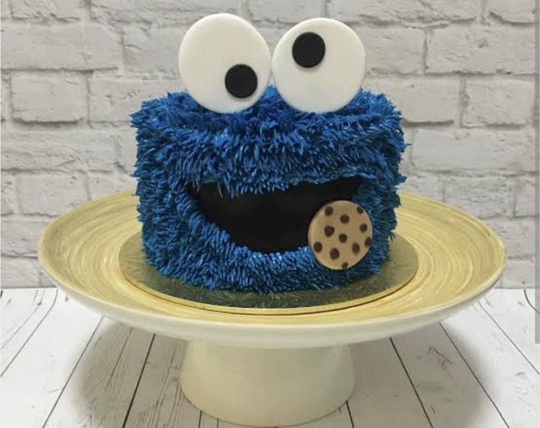 کیک به شکل هیولایی پشمالو با چشم‌های بزرگ که یک کوکی در دست دارد، روی بشقاب و پس‌زمینهٔ دیوار آجری