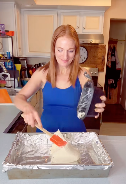 Person in a kitchen spreading dough on a foil-lined tray, with a plastic covering in hand. Kitchen items are visible in the background