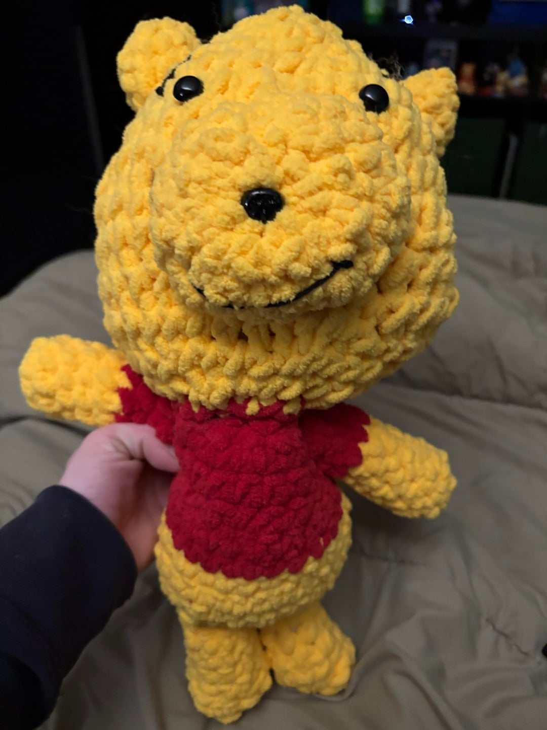 اسباب‌بازی خرسی نرم با بافت بافتنی، پیراهن قرمز و چهره‌ای خندان، توسط کسی در رختخواب نگه داشته شده