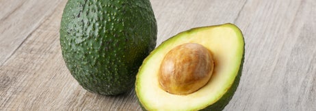木のテーブルに置かれたアボカド。1つは皮付き、もう1つは半分に切られて種が見えています。
