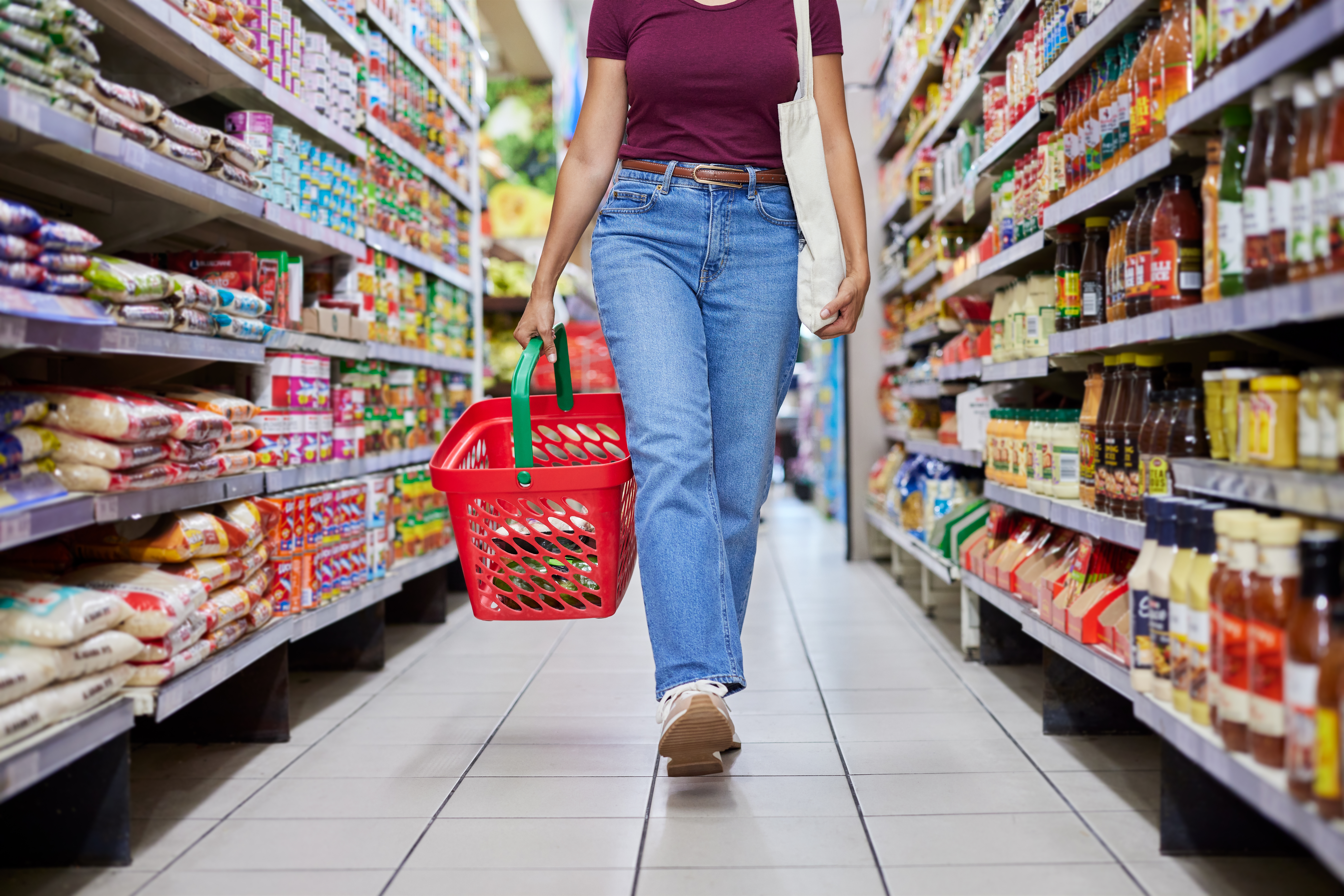 Person walking in a grocery aisle holding a basket and a tote bag, surrounded by various food products on shelves
