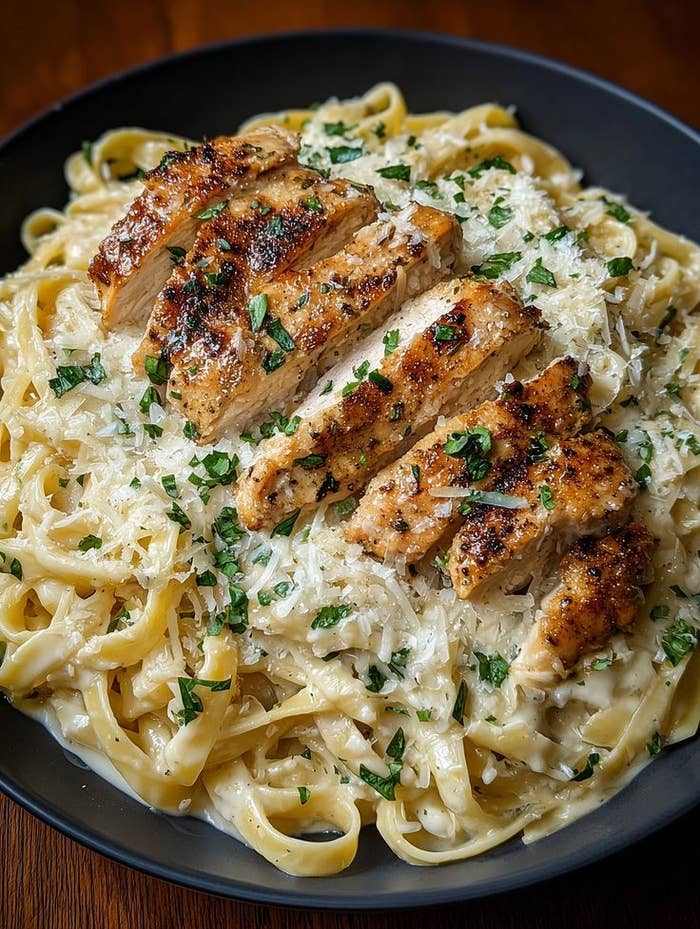 Fettuccine Alfredo con pollo a la parrilla, cubierto con queso rallado y perejil, servido en un plato oscuro